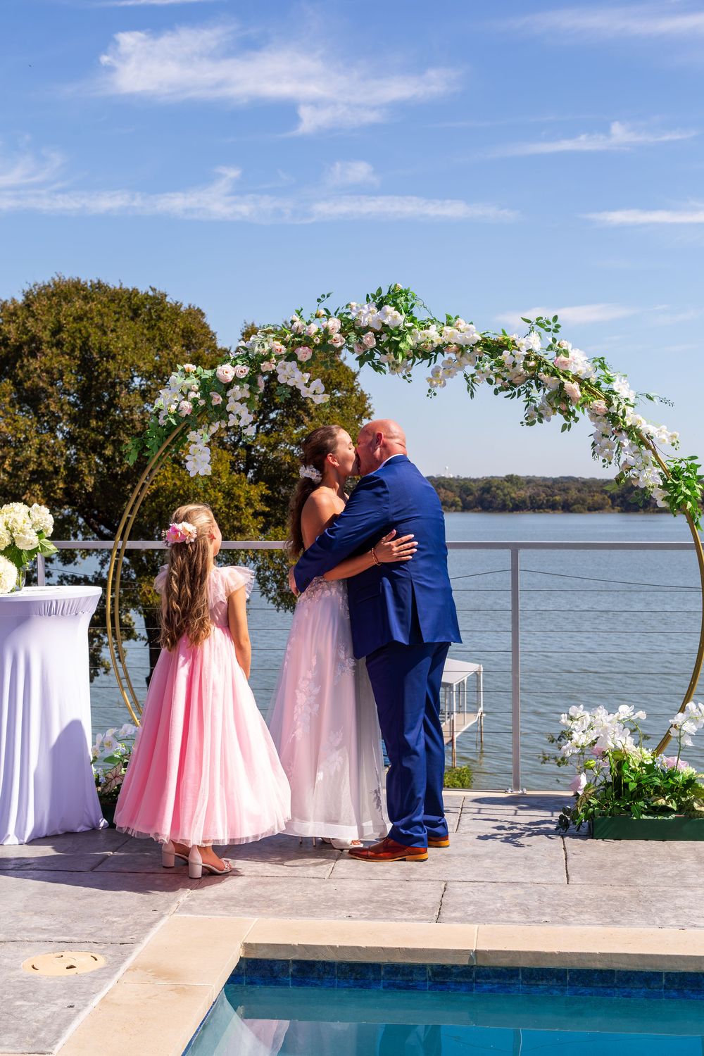 Bride and groom share first kiss under floral arch by waterfront pool with flower girl in pink dress.