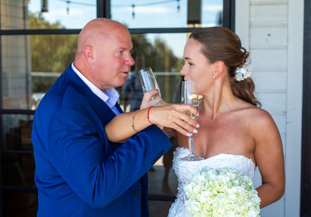 Bride and groom toasting with champagne glasses at wedding, bride holding white floral bouquet outdoors.