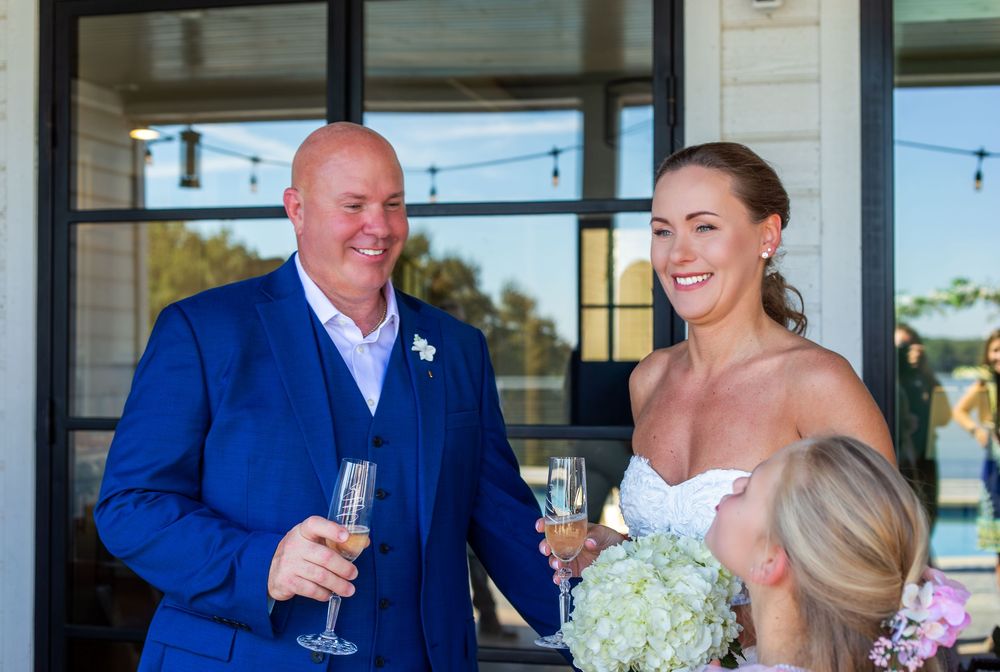 Bride in white gown and groom in blue suit toasting with champagne at outdoor wedding reception.