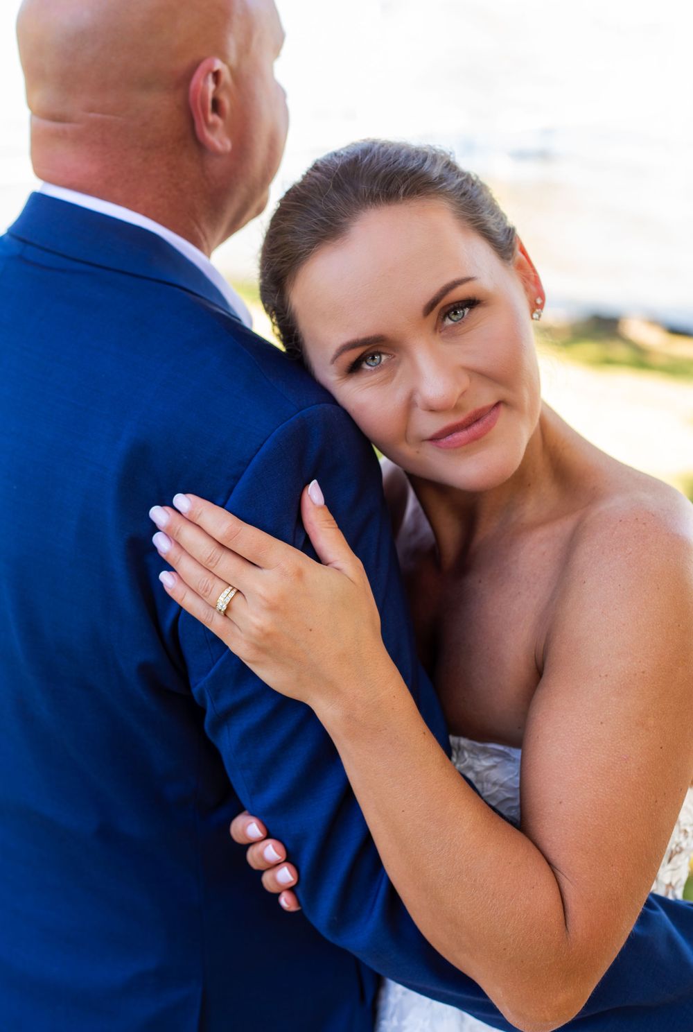 Bride in strapless gown embracing groom in navy suit, showing wedding ring, with ocean backdrop.