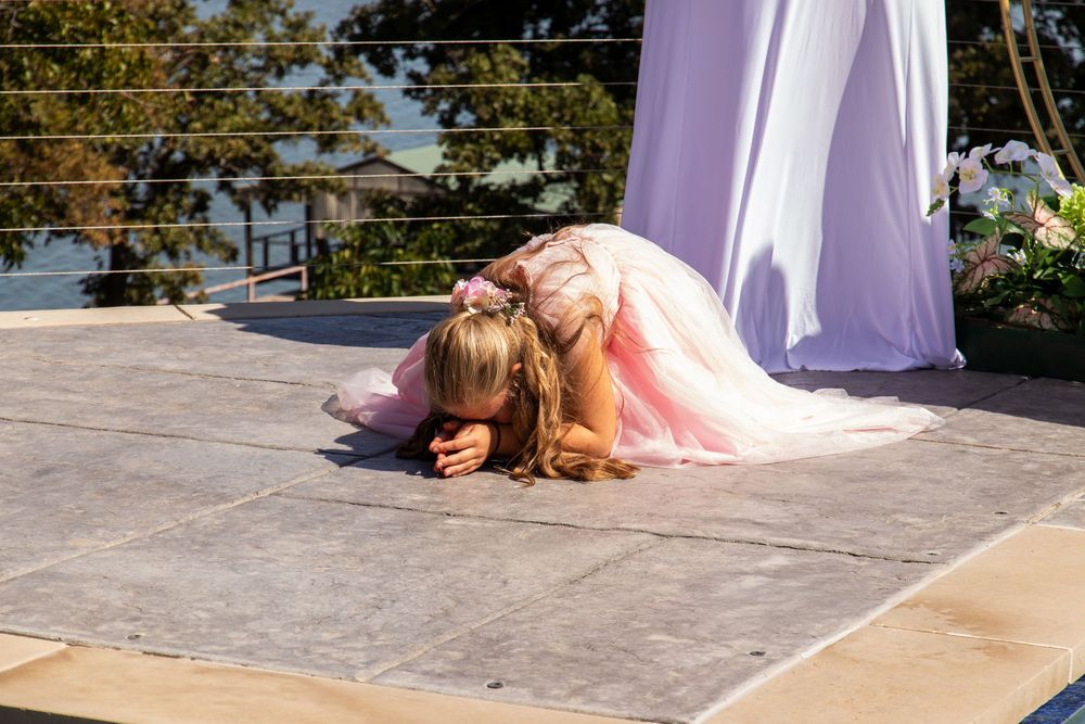 Young flower girl in pink dress bowing on outdoor wedding ceremony stage in bright sunlight.