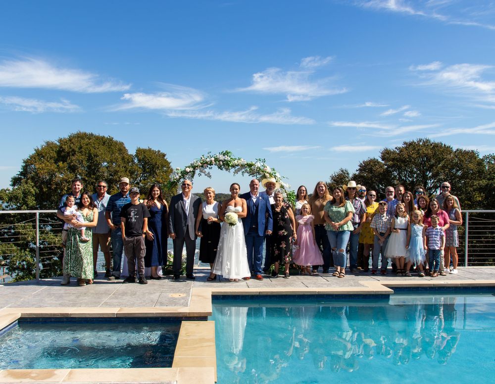 Wedding party poses poolside under floral arch with blue sky backdrop at elegant outdoor venue.