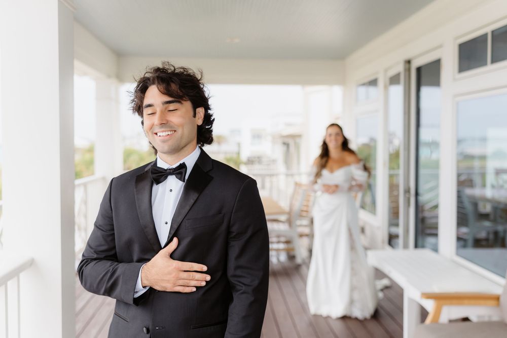 Groom in black tuxedo smiles with hand on chest during first look, bride in white gown stands in background on porch.