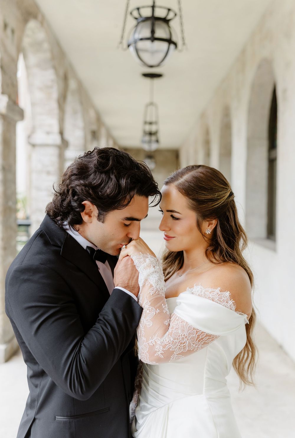 Groom in black tuxedo kissing bride's hand in lace off-shoulder wedding dress under elegant archway.