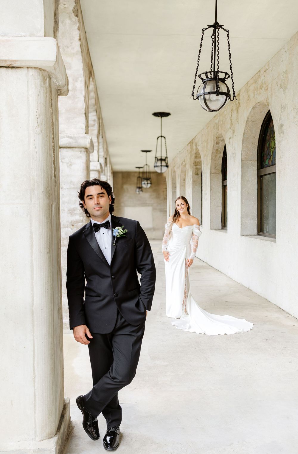 Groom in black tuxedo stands in arched corridor as bride in white gown approaches from behind.