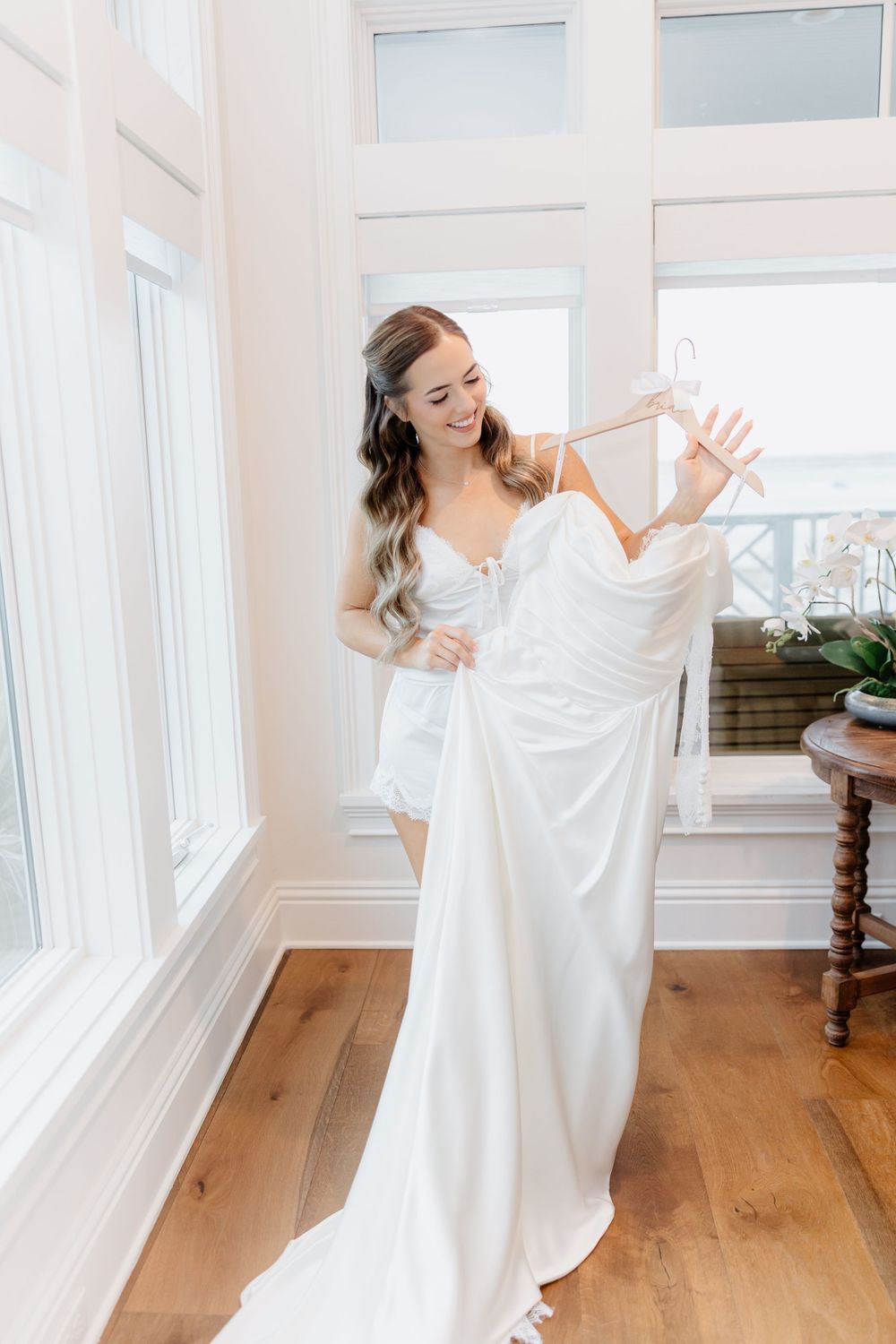 Bride in white slip dress holds her elegant wedding gown on a hanger near bright windows in a sunlit room.
