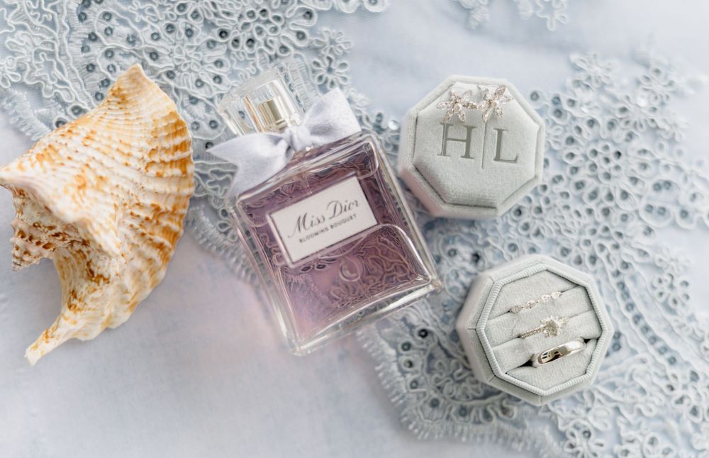 Miss Dior perfume bottle beside a velvet ring box with HL monogram, wedding rings, and a seashell on lace.