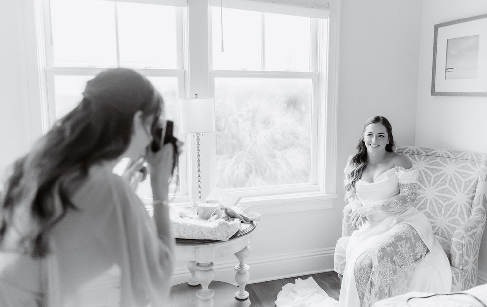 Bride in white gown sits by window while photographer captures her portrait in elegant black and white wedding photo.