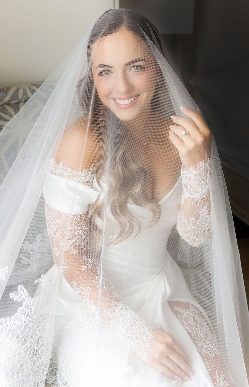 Smiling bride in off-shoulder lace wedding gown and veil poses elegantly indoors.