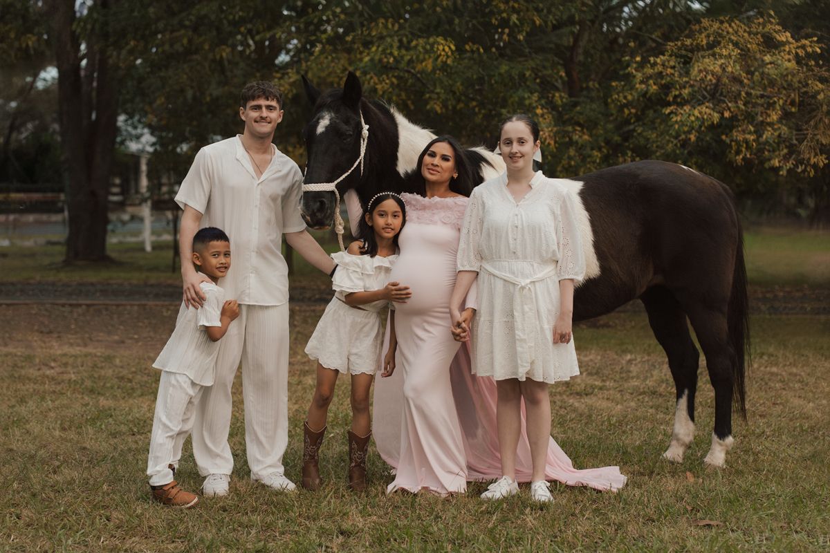 Family posing outdoors with a black and white horse, wearing coordinated white and pink outfits. Family photoshoot with horses in sydney