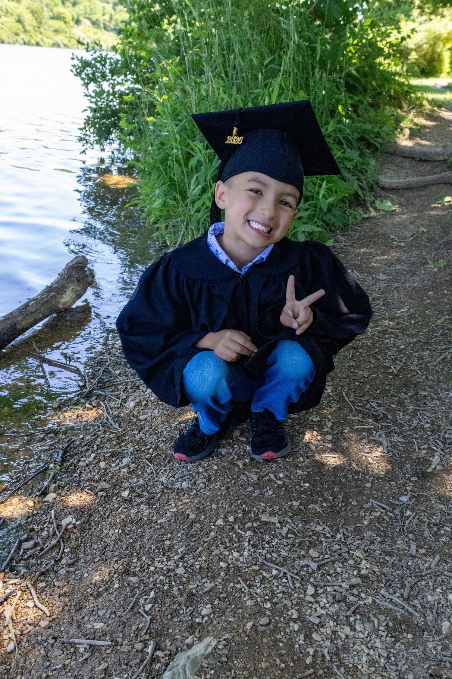 Smiling boy in graduation cap and gown crouching by a lake, making a peace sign outdoors.