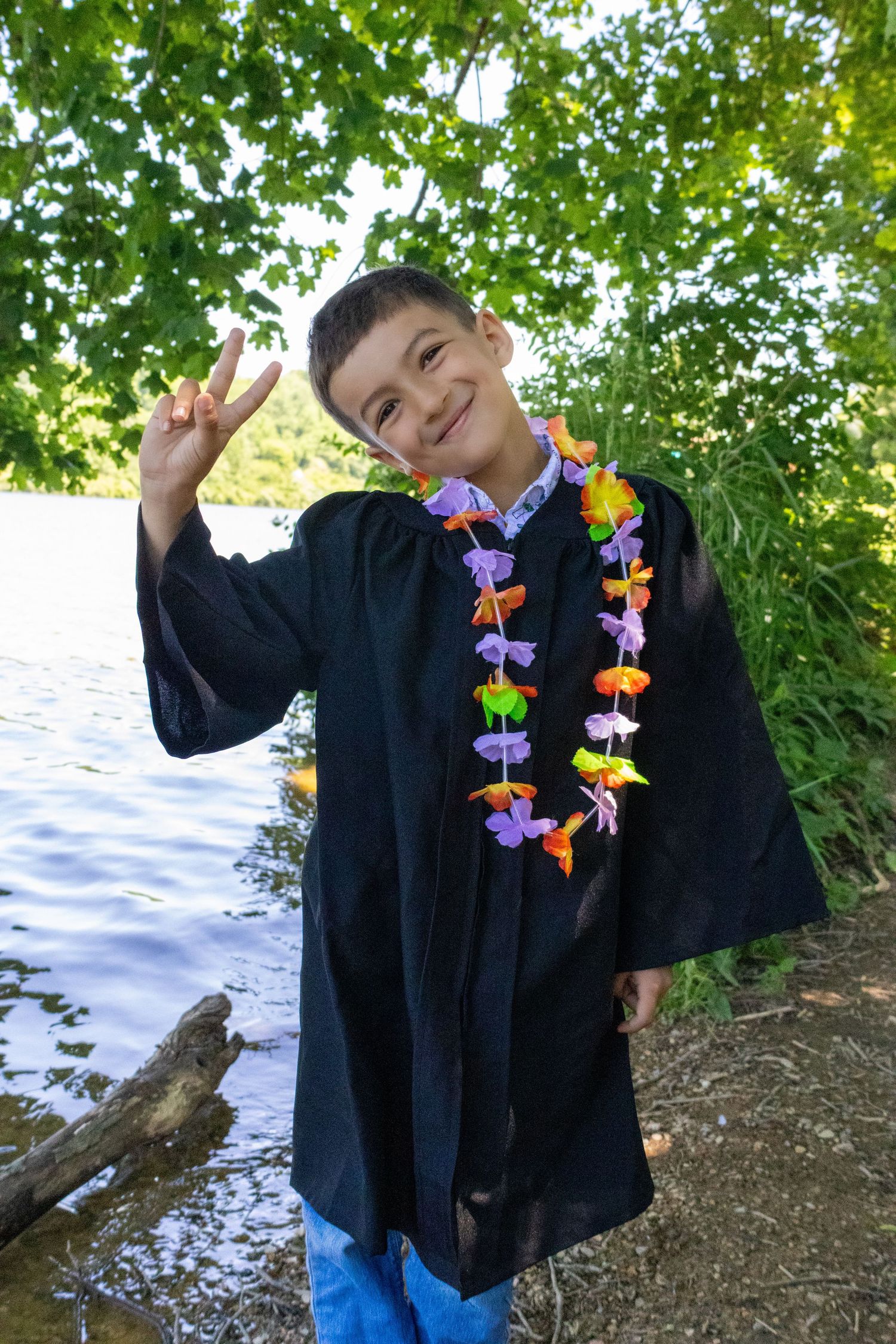 Smiling boy in black graduation gown and colorful lei making peace sign by a lake with green trees.