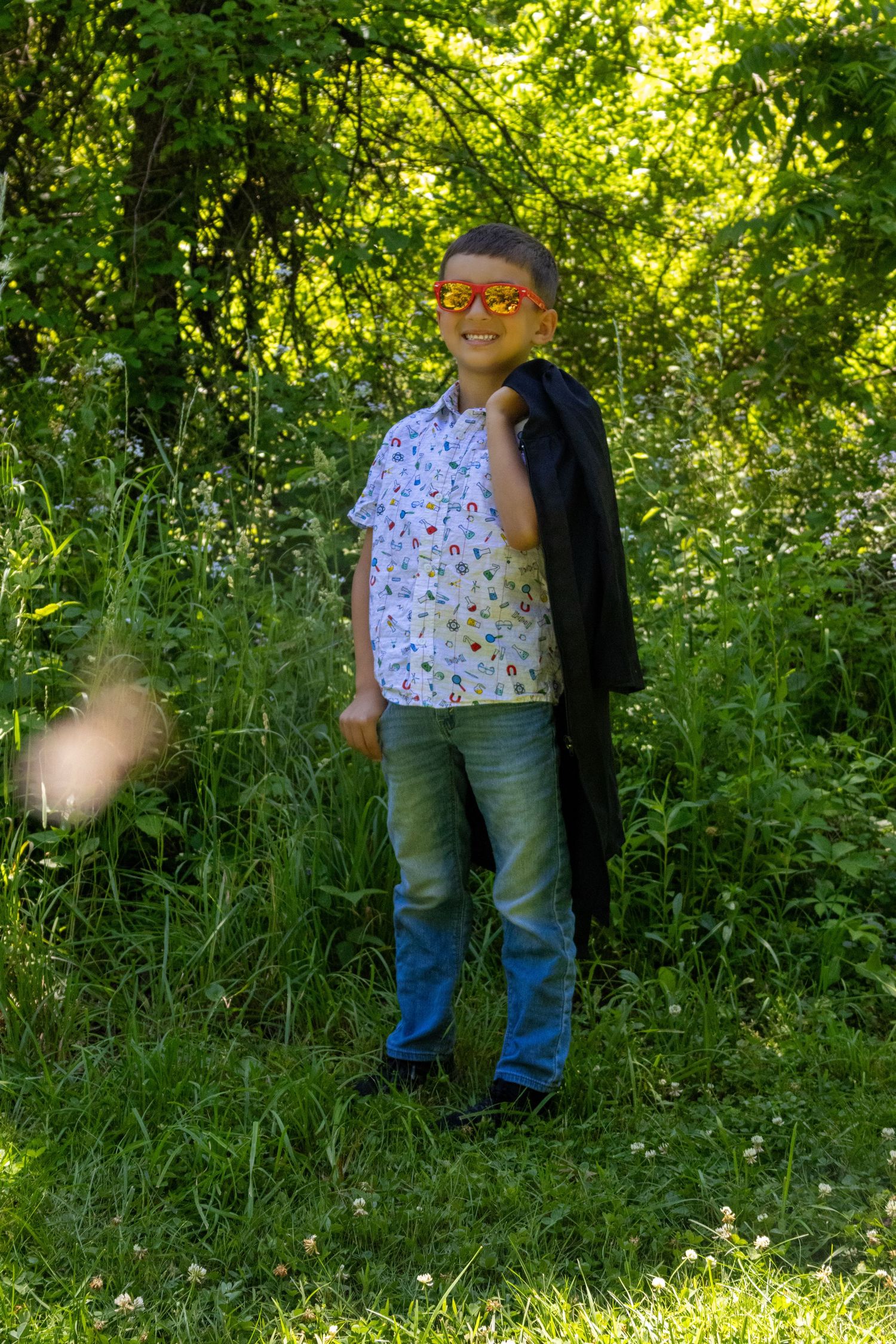 Young boy dressed as a superhero with orange mask and black cape smiling outdoors in lush green nature.