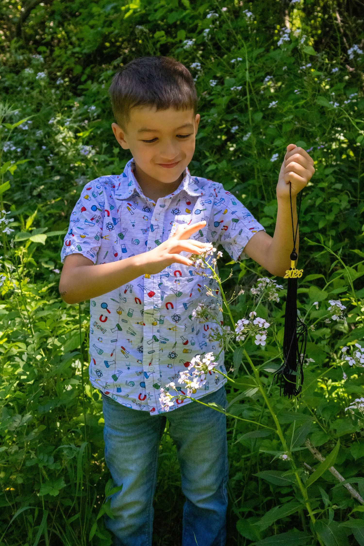 A young boy in a patterned shirt holds a graduation tassel while standing among wildflowers and green foliage outdoors.