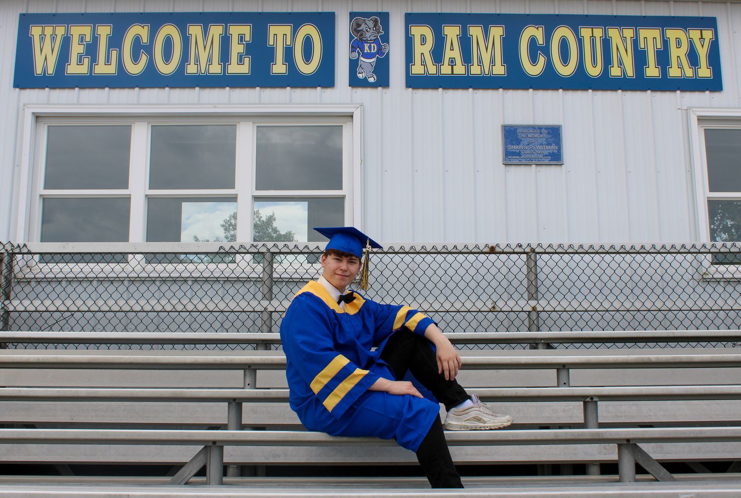 Graduate in blue and gold cap and gown sits on bleachers in front of a Welcome to Ram Country sign.