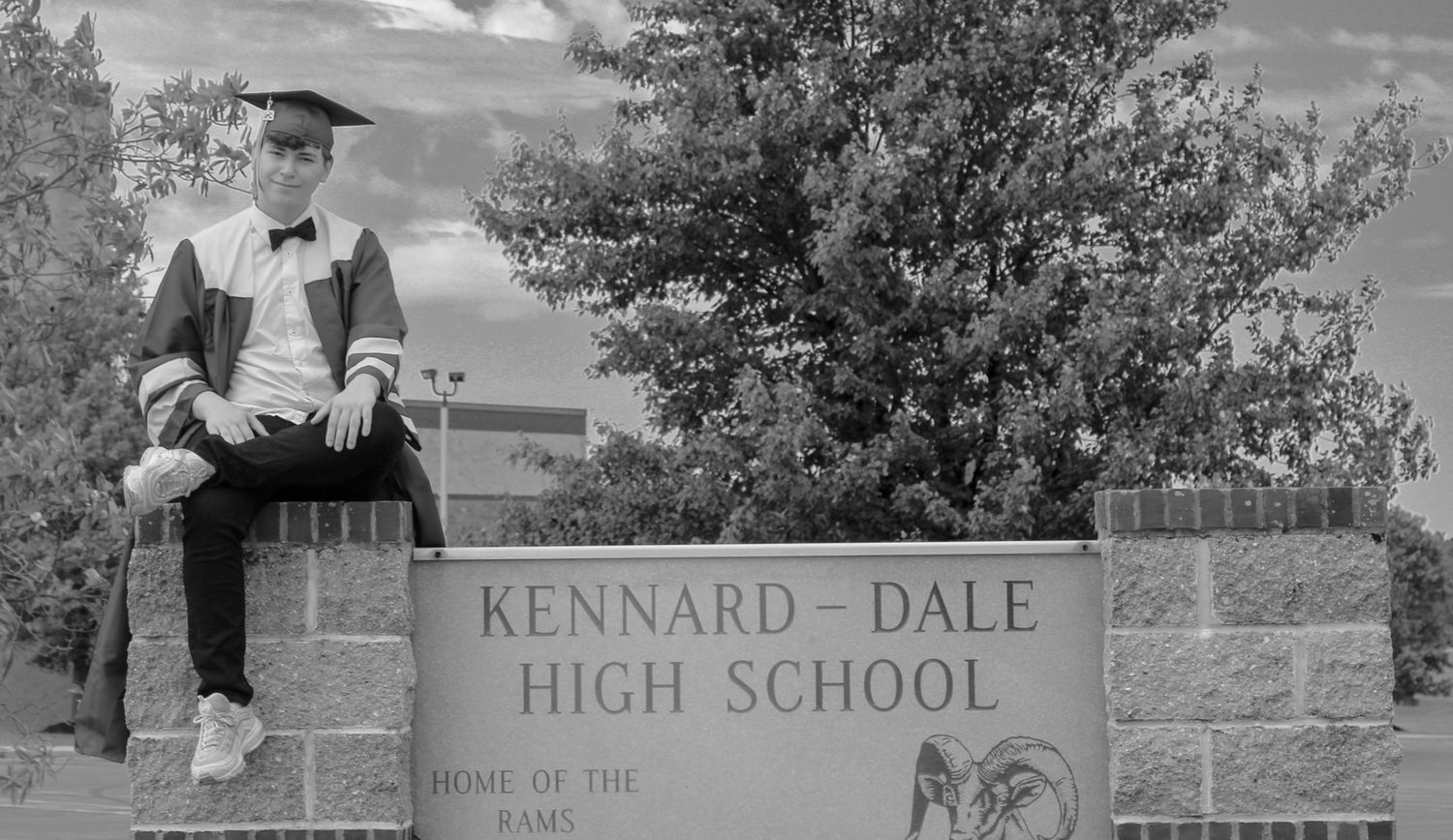 Graduate in cap and gown poses beside Kennard-Dale High School sign, Home of the Rams.