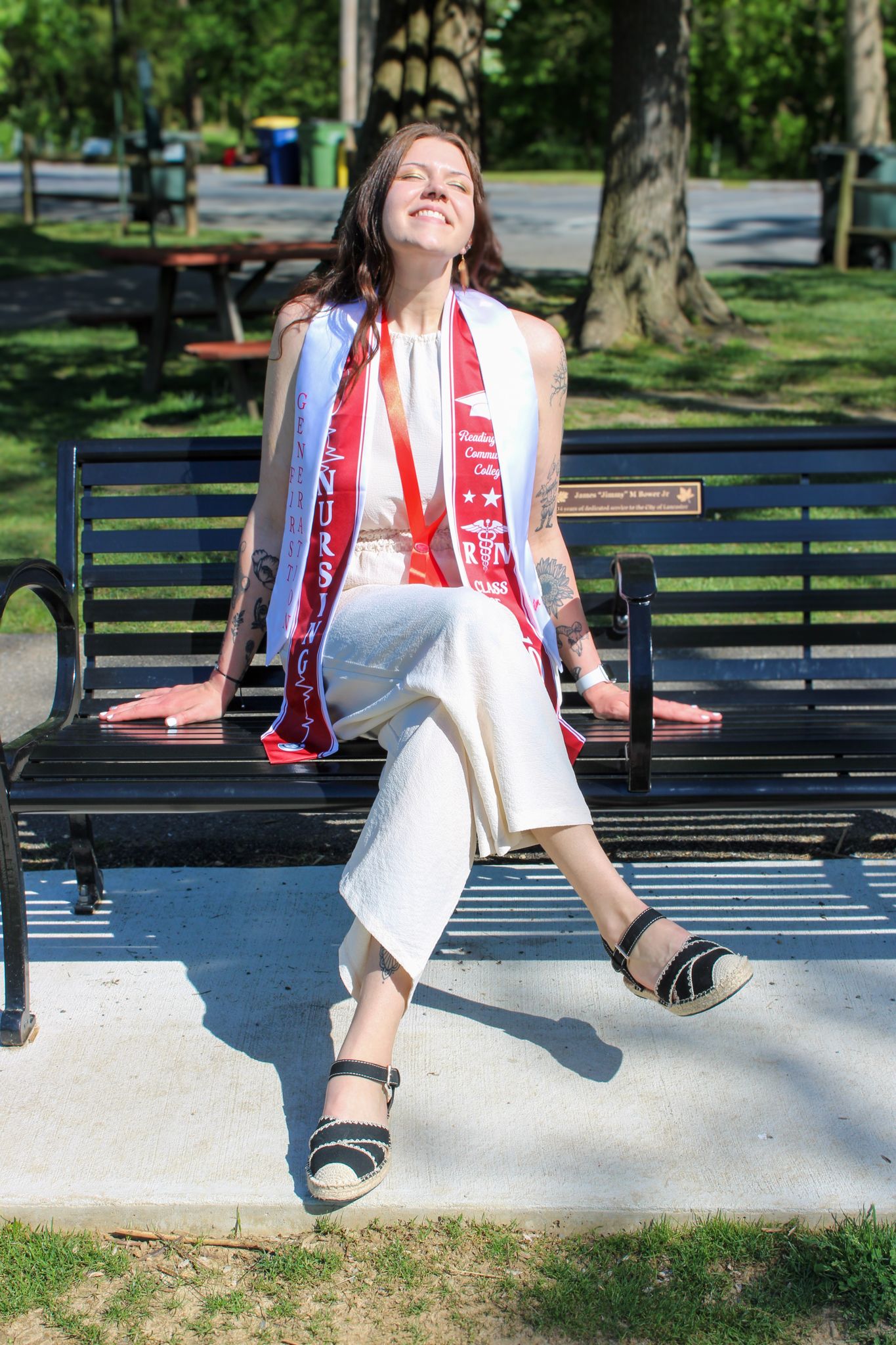 Smiling graduate sits on park bench wearing white outfit with red graduation stoles and medals on sunny day.