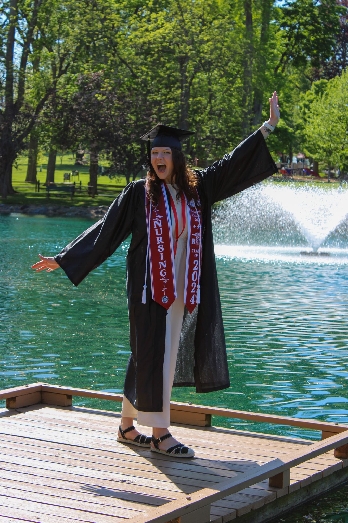 Nursing graduate in cap and gown celebrates by fountain, arms raised joyfully on wooden dock surrounded by green trees.