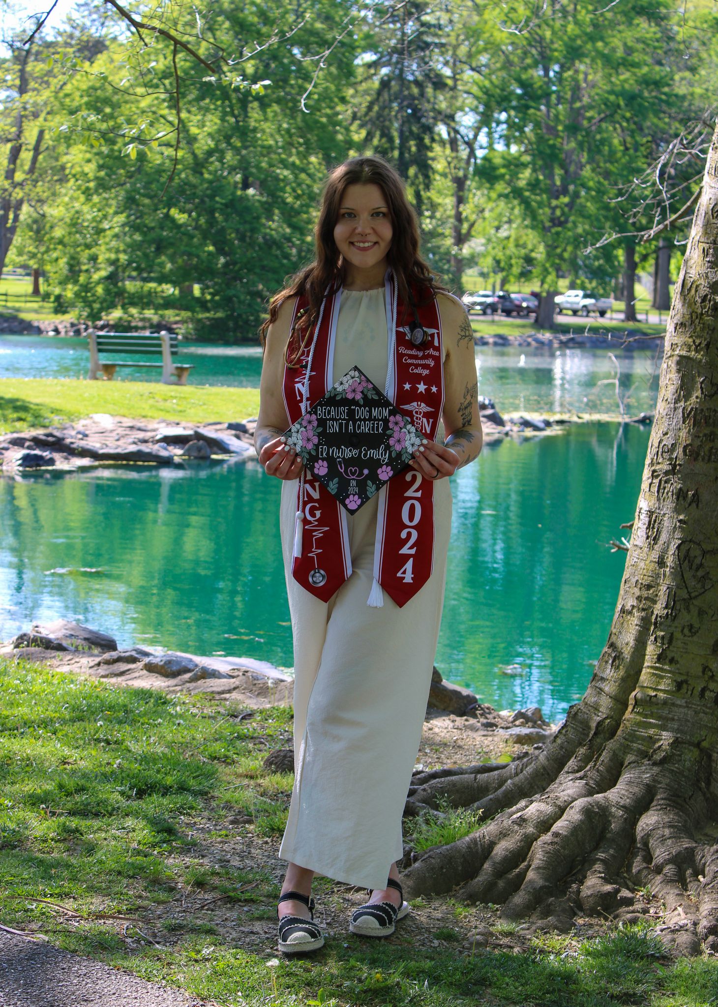 A smiling 2024 graduate in a white dress wearing red graduation stoles poses by a scenic pond in a park.