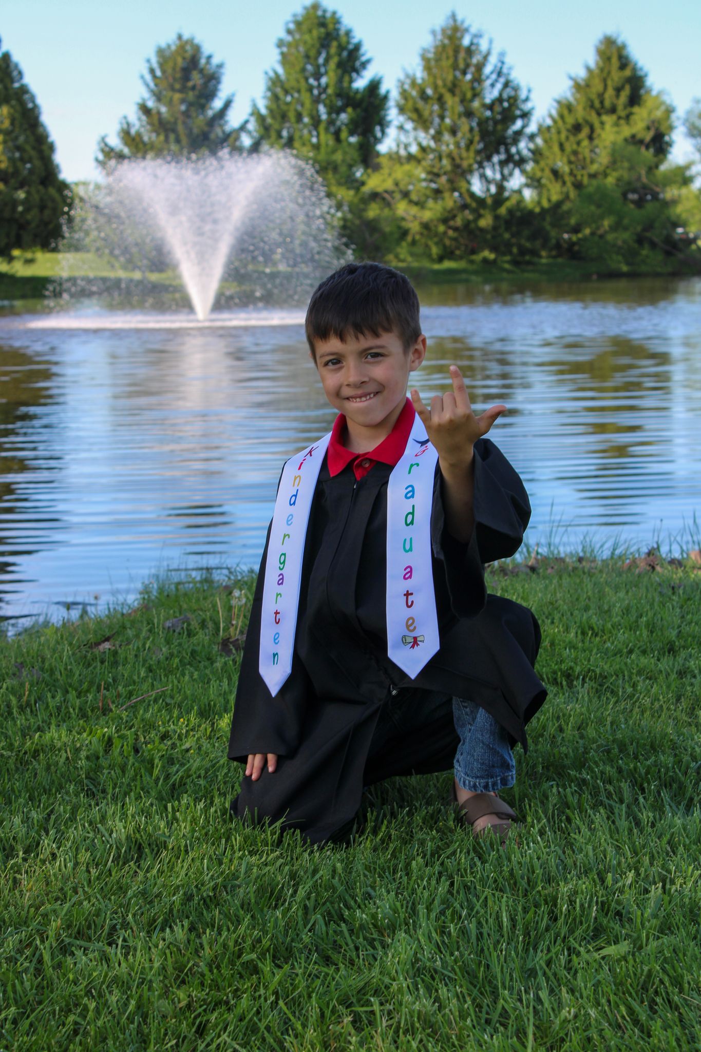 Young boy in graduation gown and sash kneeling on grass near fountain, making shaka sign, celebrating kindergarten graduation.