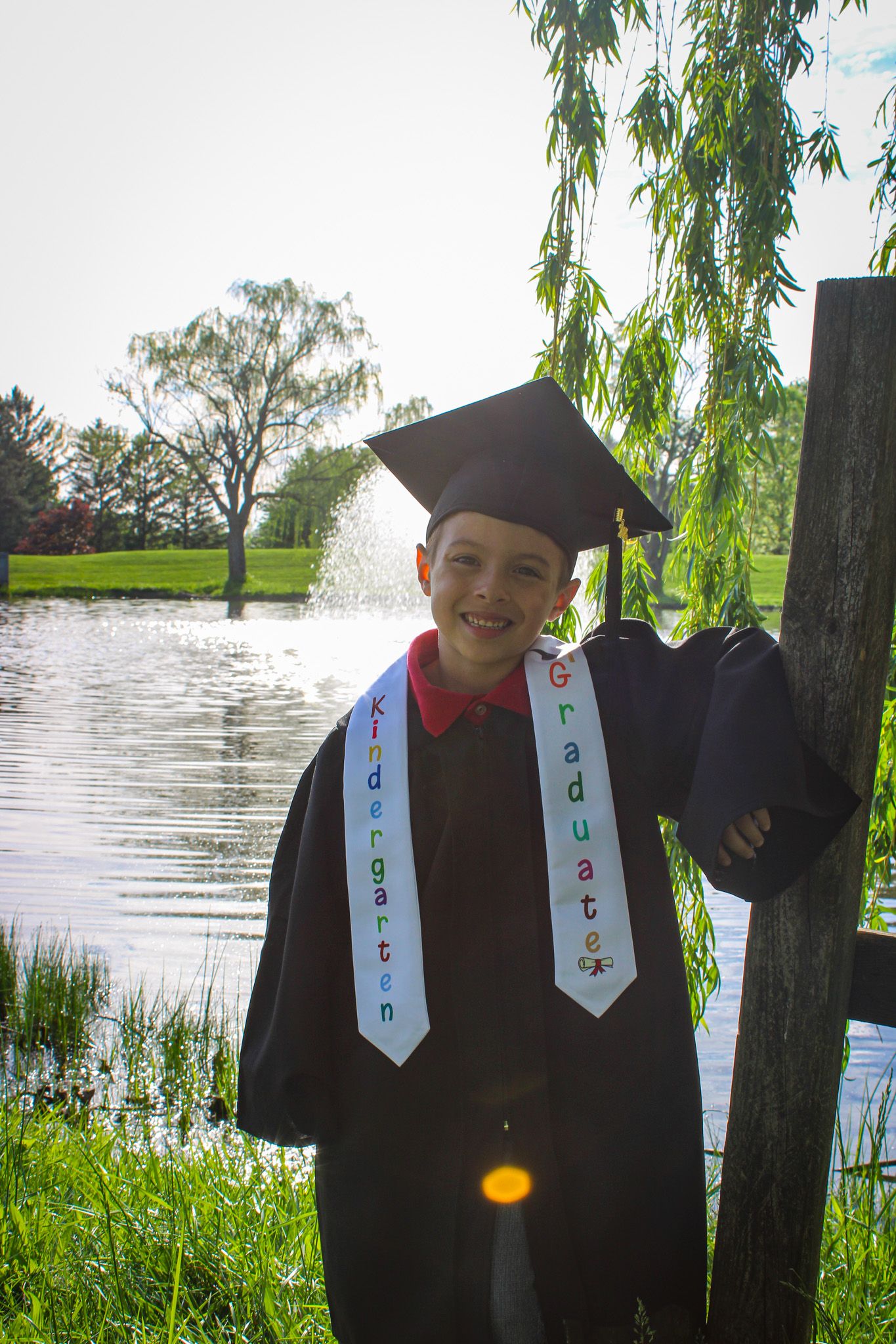 Young boy in graduation cap and gown with Kindergarten Graduate sash smiling outdoors near a pond.