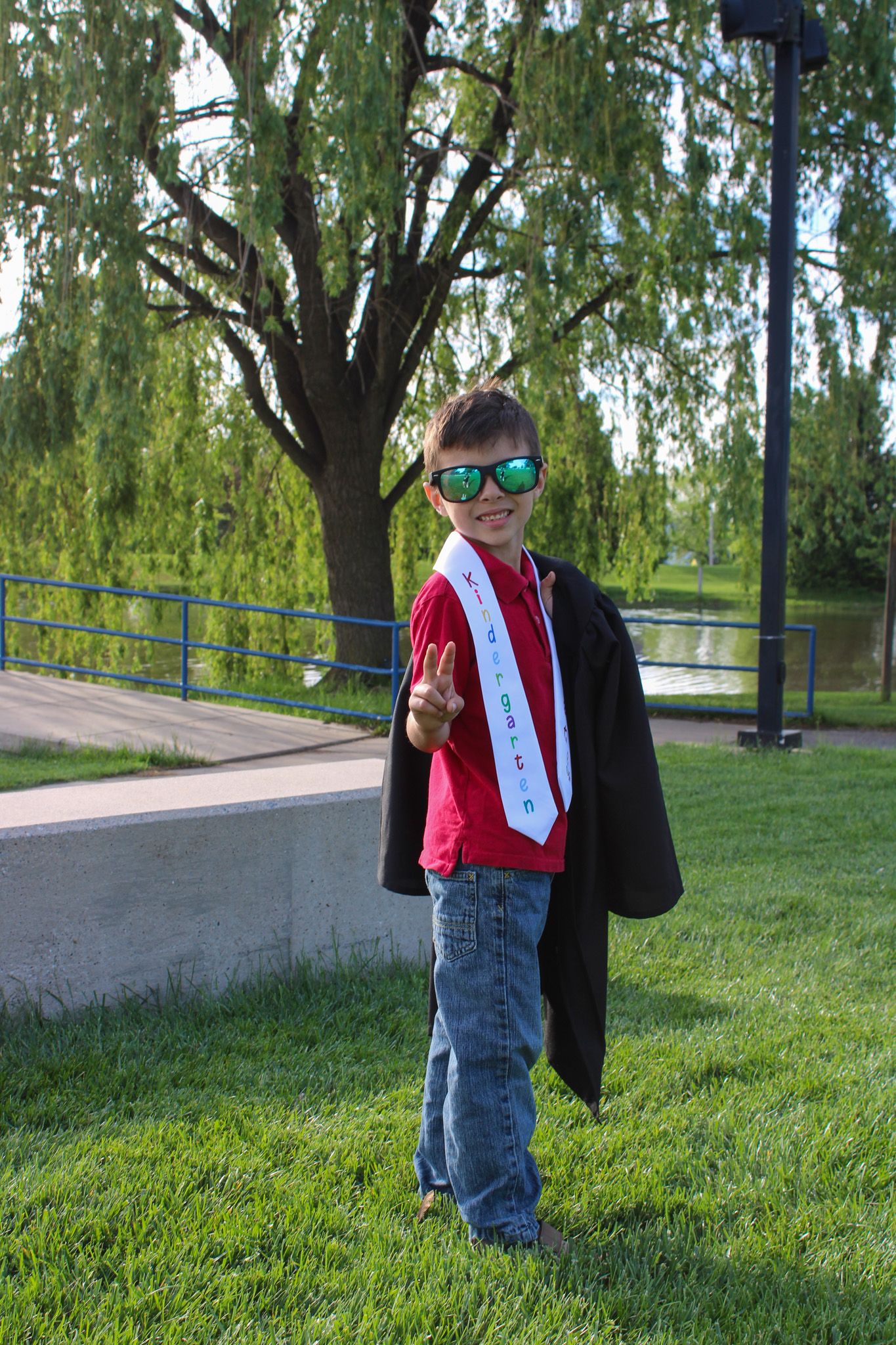 Young boy in graduation cap and gown with green sunglasses giving peace sign outdoors near willow tree.