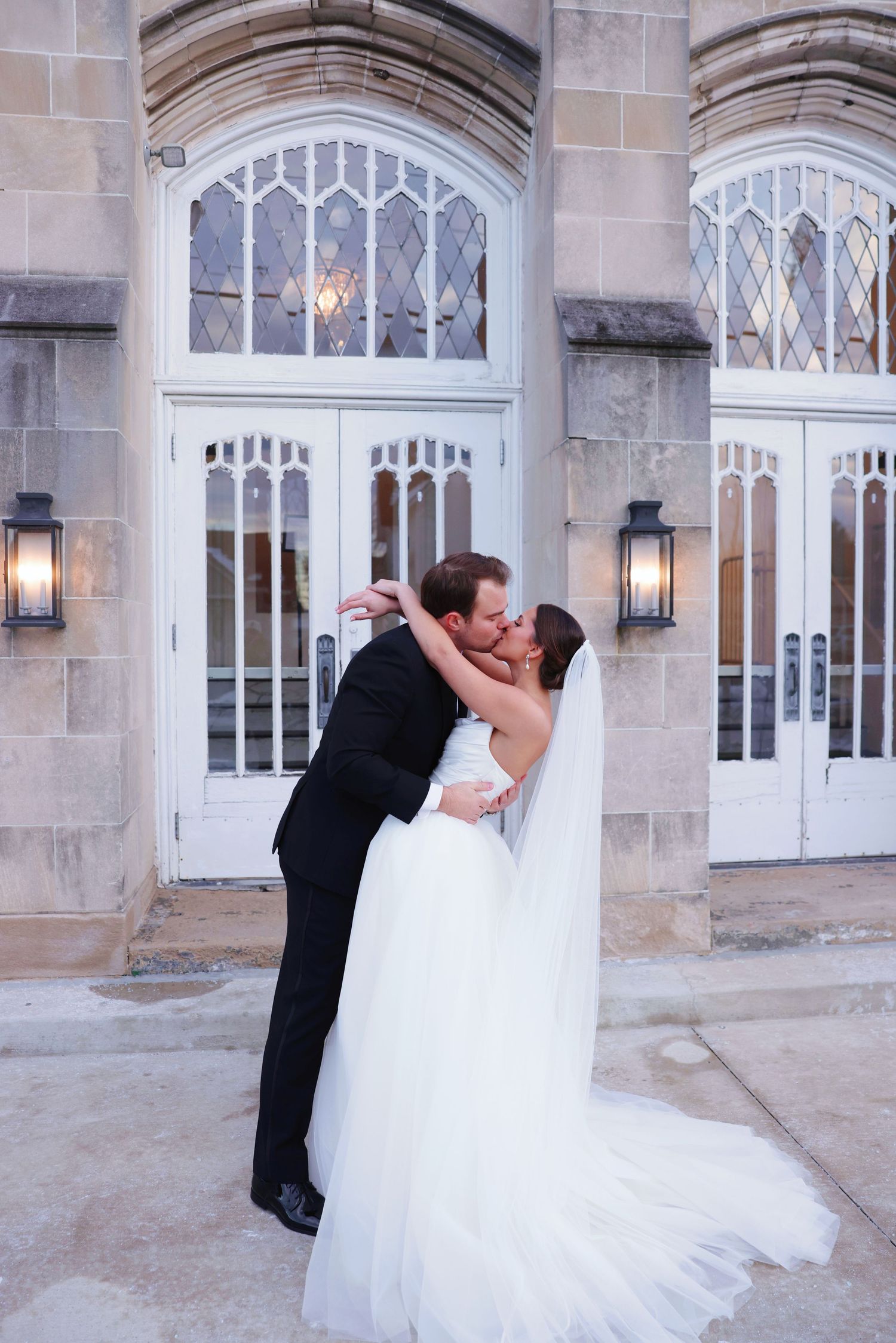 Bride and groom share a romantic kiss outside a stone church, bride in white gown with veil trailing behind her.