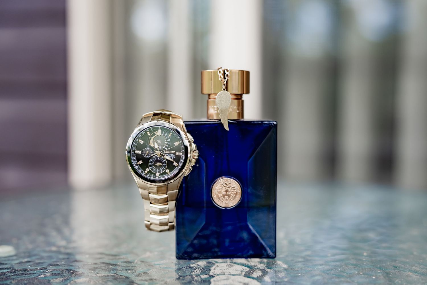 Luxury gold chronograph watch beside a blue Versace cologne bottle on a reflective glass surface.
