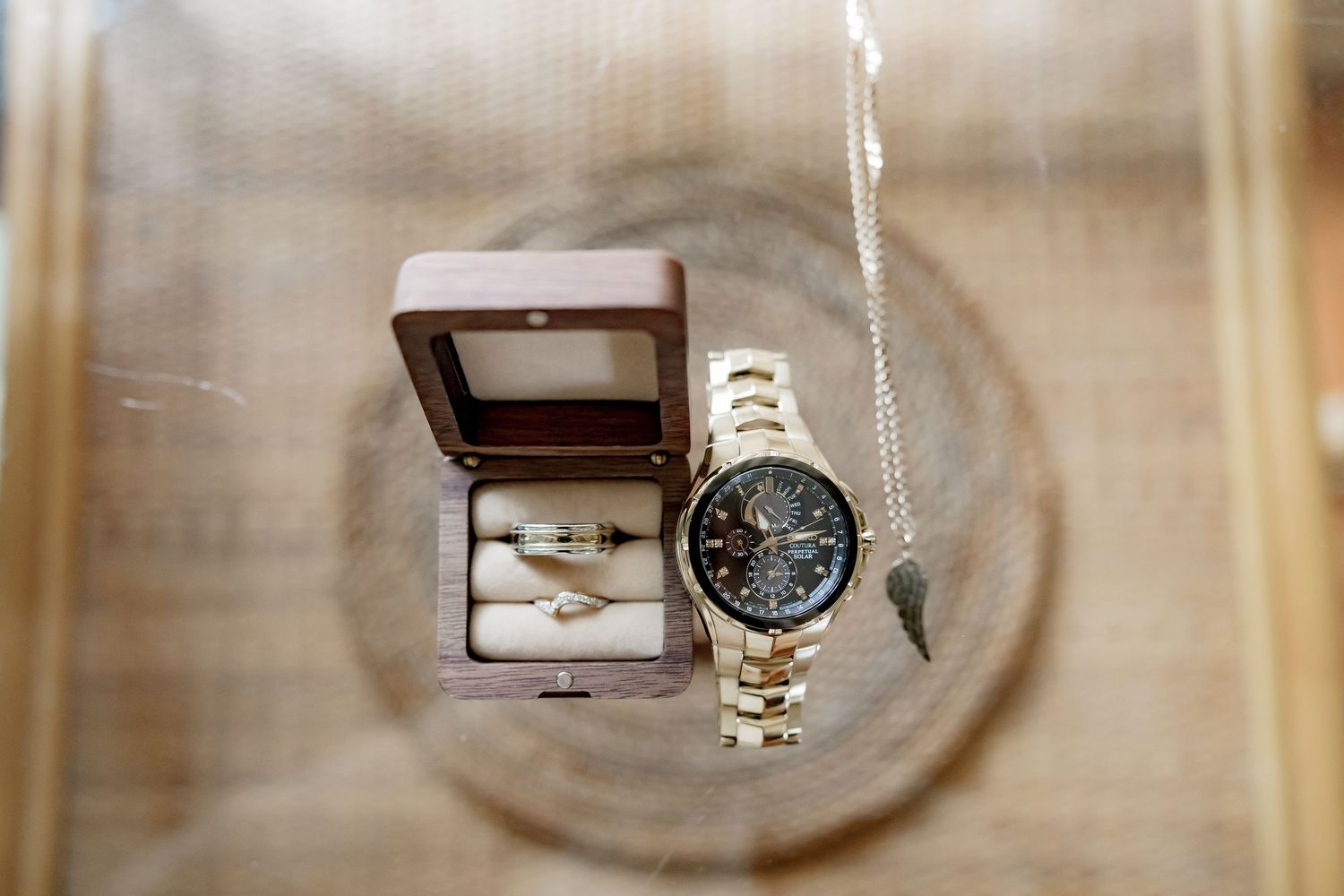 Gold watch, wedding rings in a velvet box, and a delicate necklace arranged on a wooden surface.