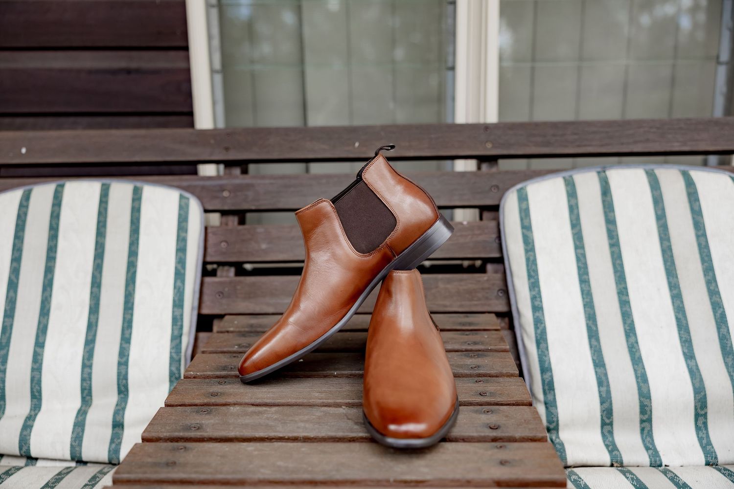 Pair of tan leather Chelsea boots placed on a wooden outdoor table between striped cushioned chairs.