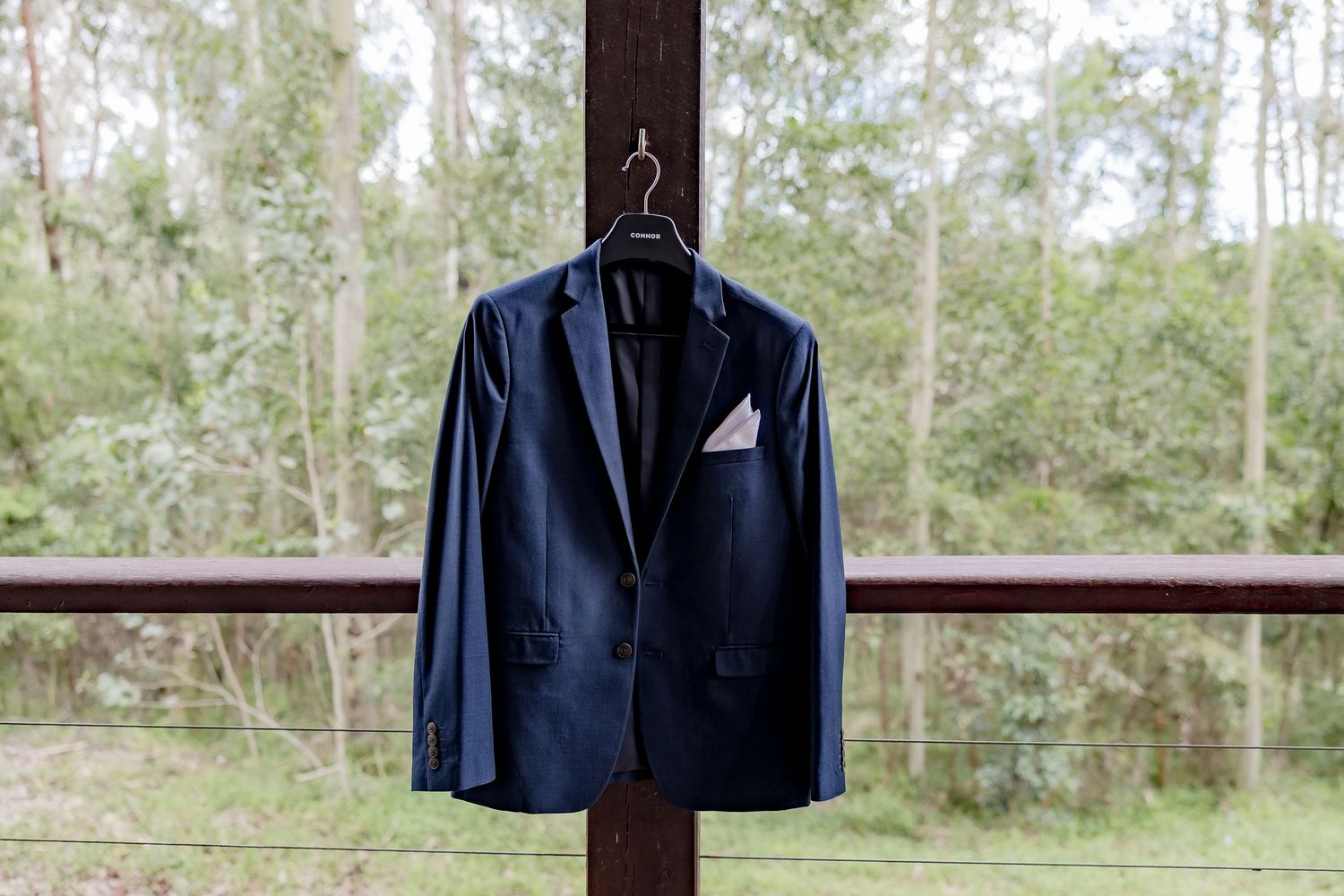 Navy blue wedding suit jacket with white pocket square hanging on wooden post outdoors.