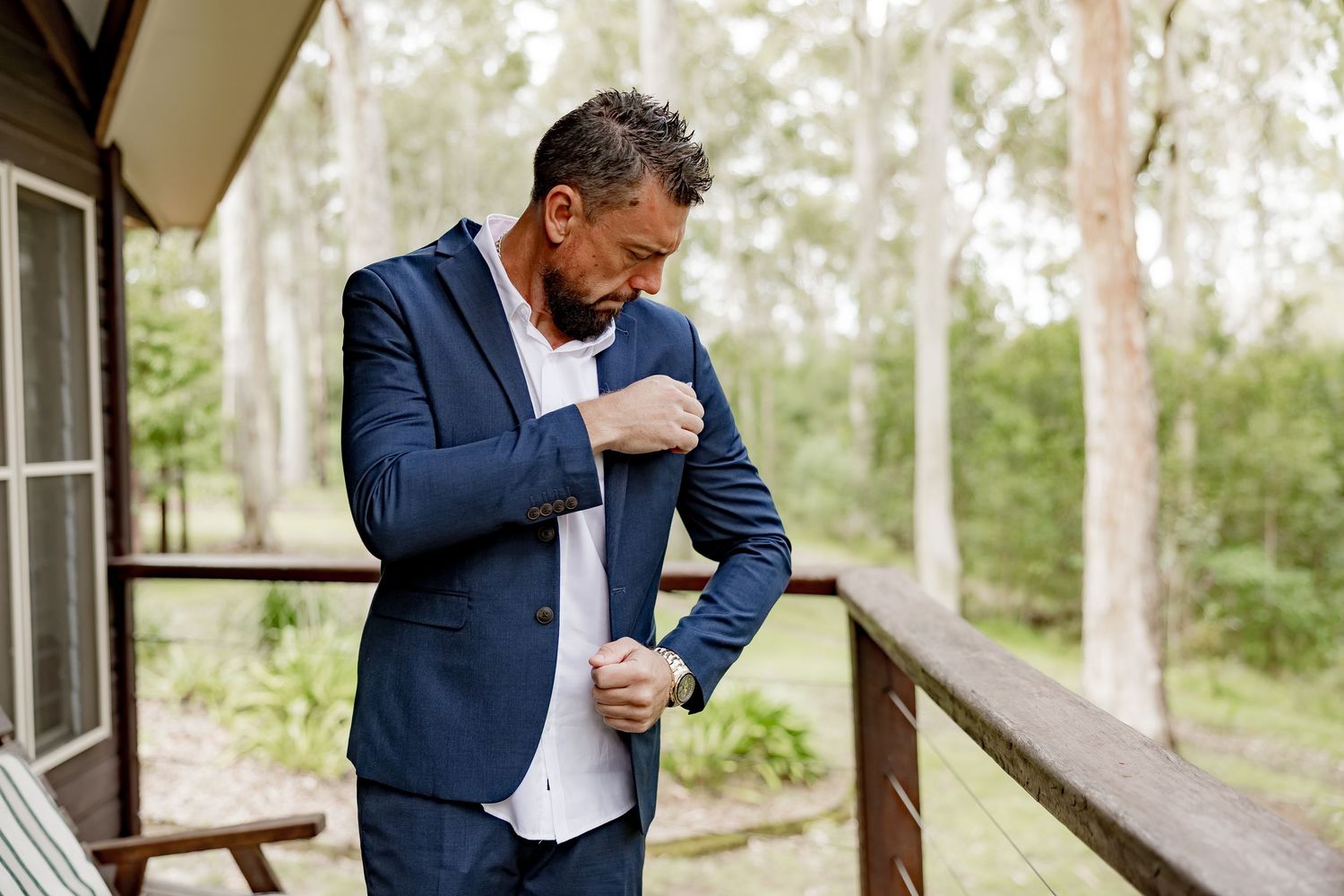 Groom in navy blue suit adjusting his jacket on a wooden deck surrounded by trees.