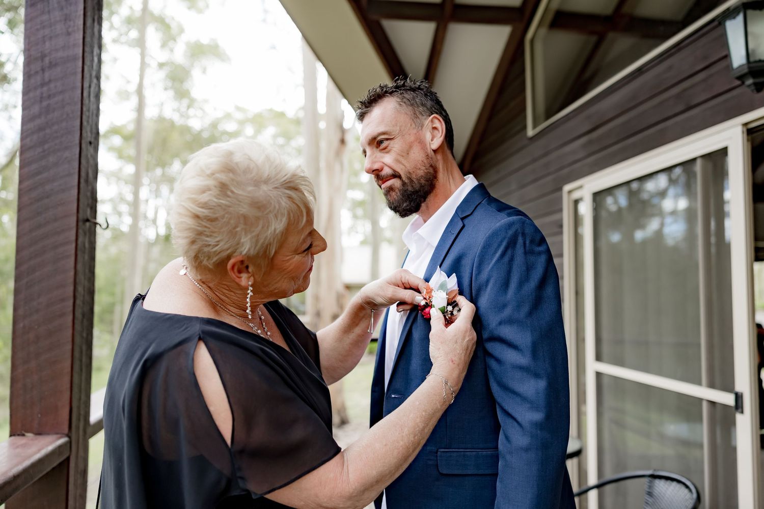 Older woman helping groom pin a boutonniere on his navy suit jacket on a wooden porch.