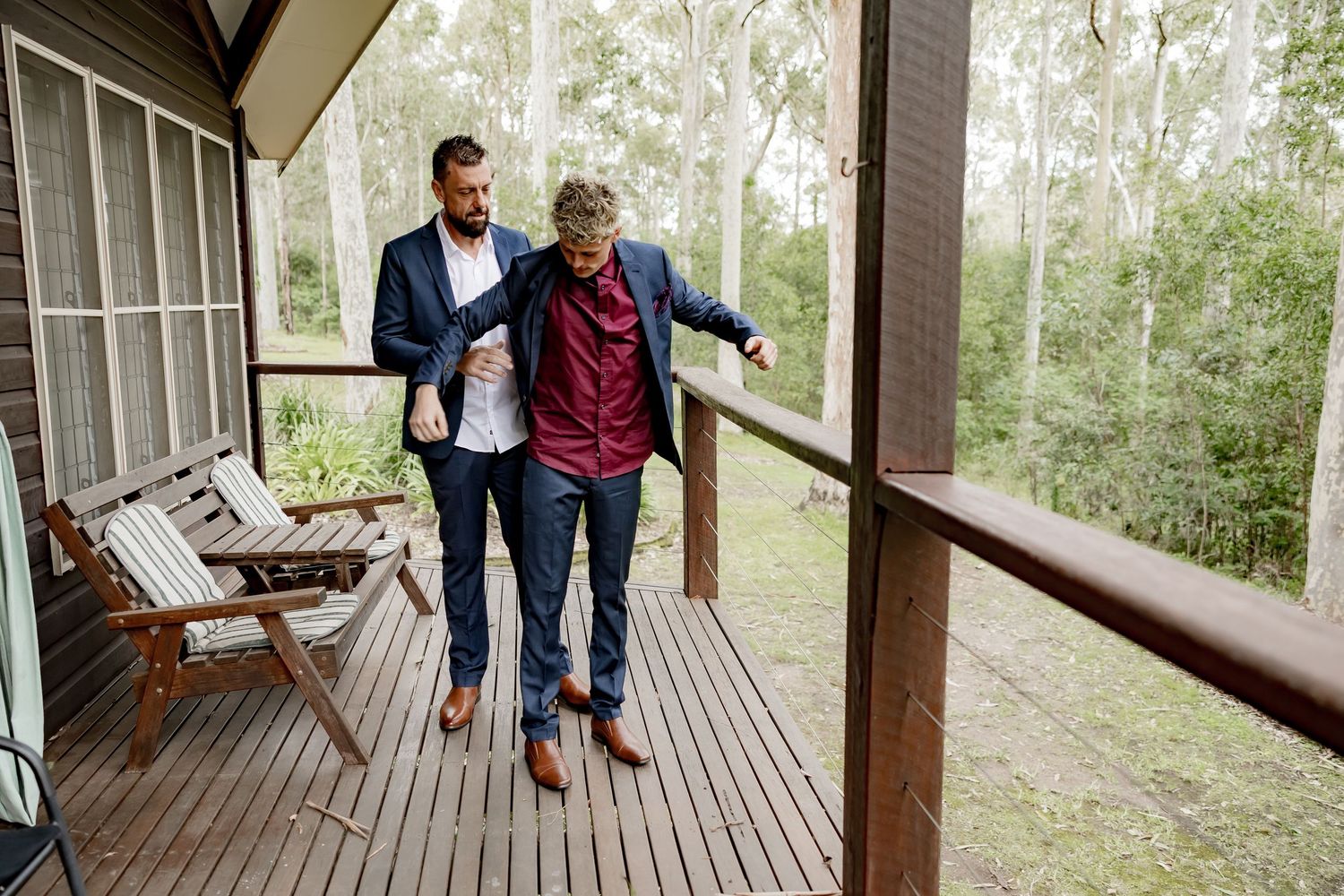 Two grooms in navy suits share a moment on a wooden deck surrounded by lush bushland.