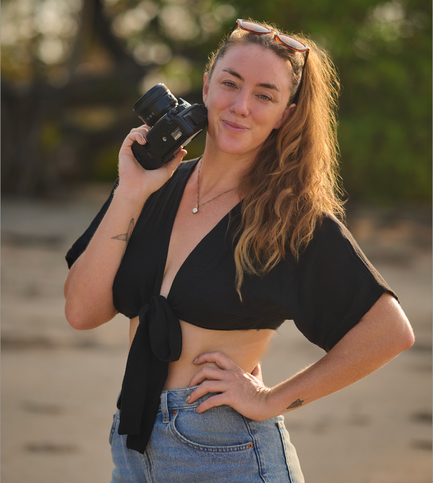 Young woman in black crop top and denim shorts holds camera on sandy beach at golden hour.