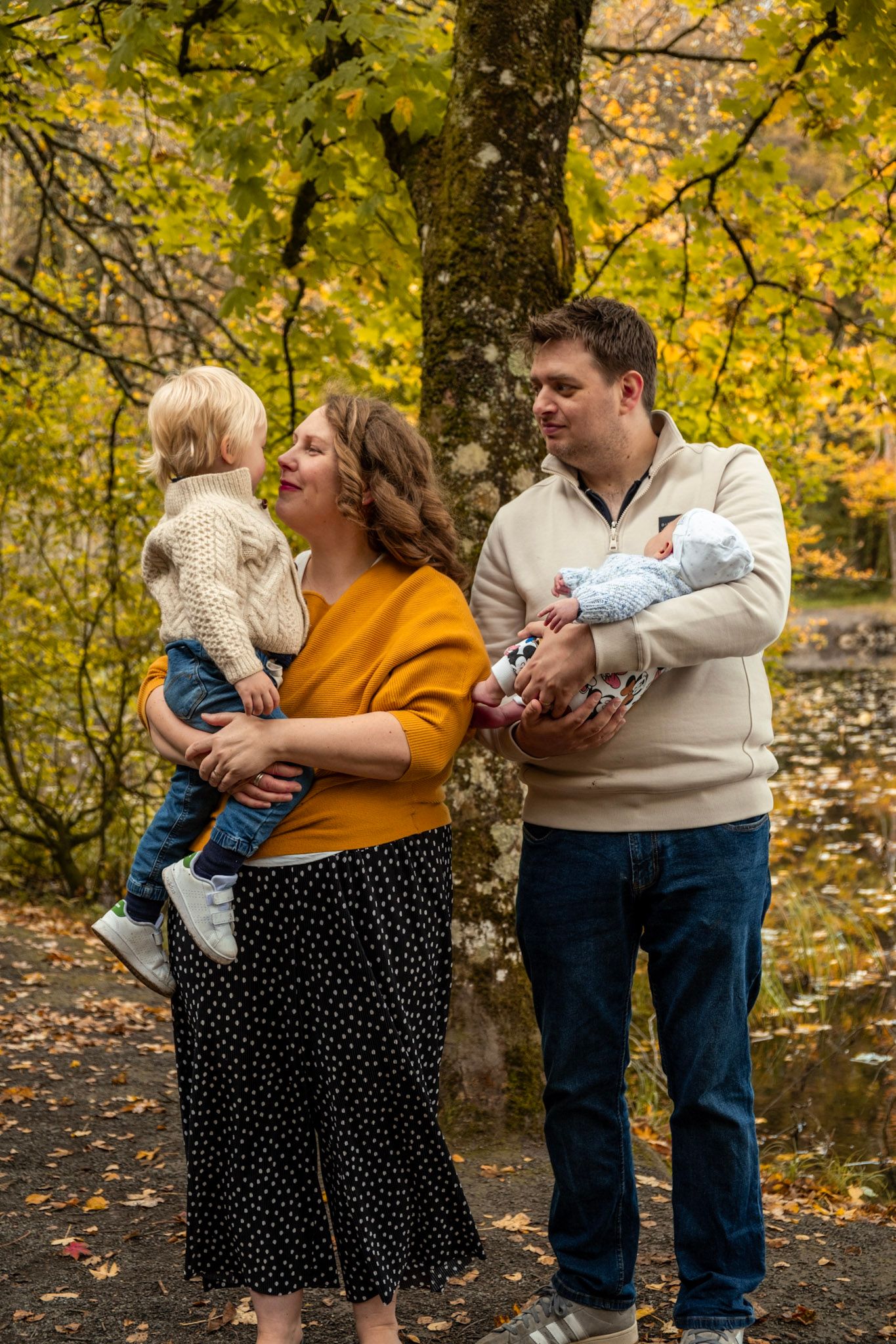 Family of four posing outdoors in autumn park, mother holding toddler, father cradling newborn baby.