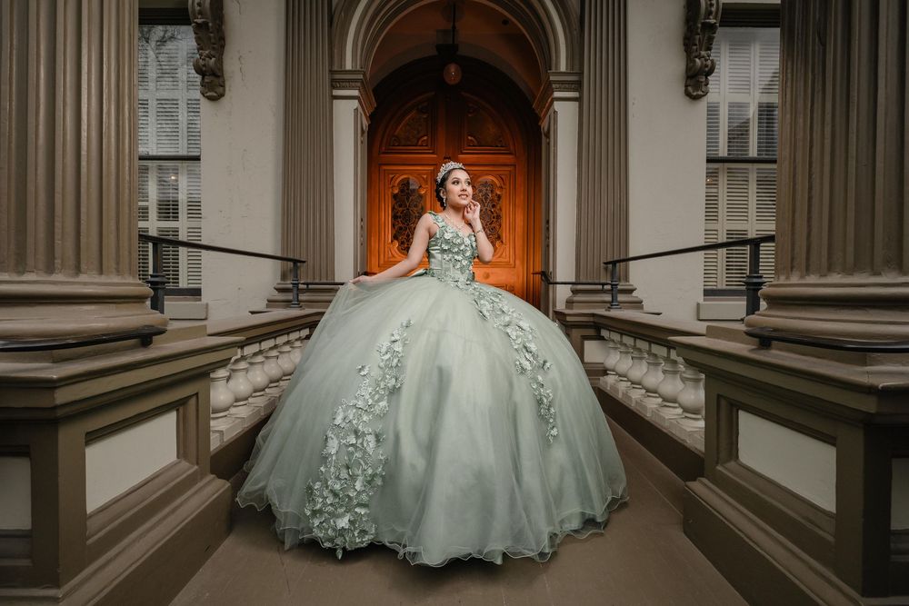 Young woman in elegant sage green ball gown with floral details poses on grand architectural steps.