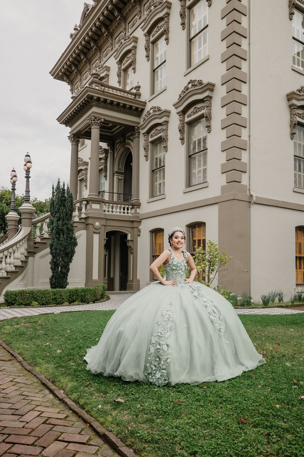 Young woman in mint green quinceañera ball gown poses in front of elegant historic mansion on green lawn.