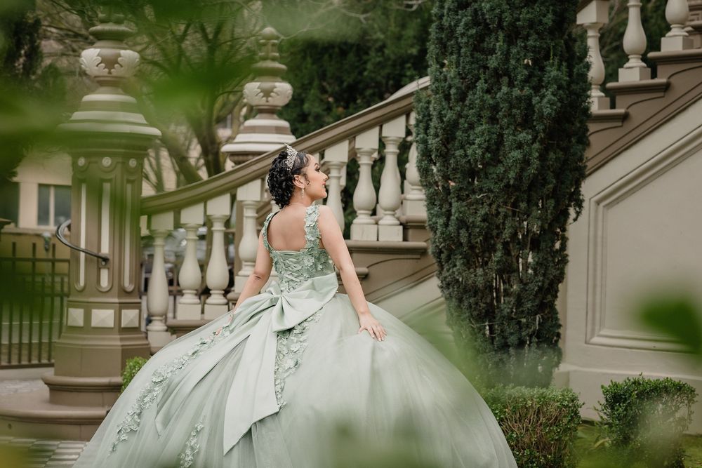 Young woman in elegant mint green quinceañera ball gown posing on ornate stone staircase surrounded by lush greenery.