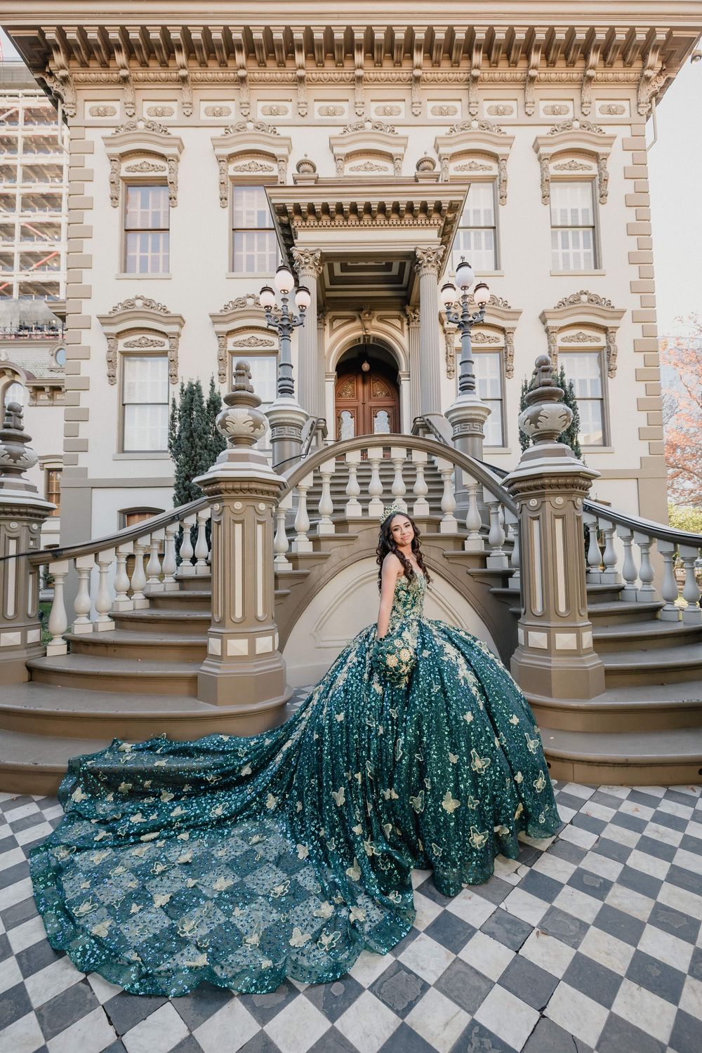 Woman in emerald green ballgown with gold details poses on grand staircase of ornate historic mansion.