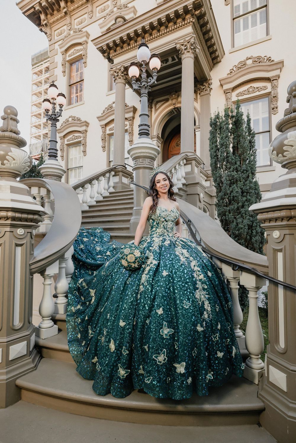 Young woman in a stunning teal ball gown with gold butterfly appliques poses on grand staircase of elegant mansion.