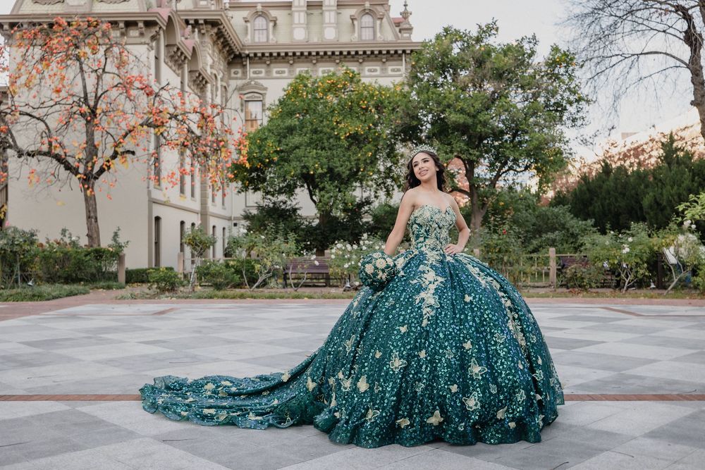 Young woman in elegant teal ball gown with floral embroidery poses in plaza for quinceañera celebration.