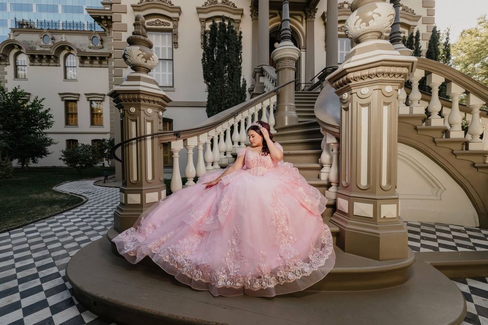 Young woman in a pink ball gown poses on grand staircase of elegant mansion for quinceañera photos.