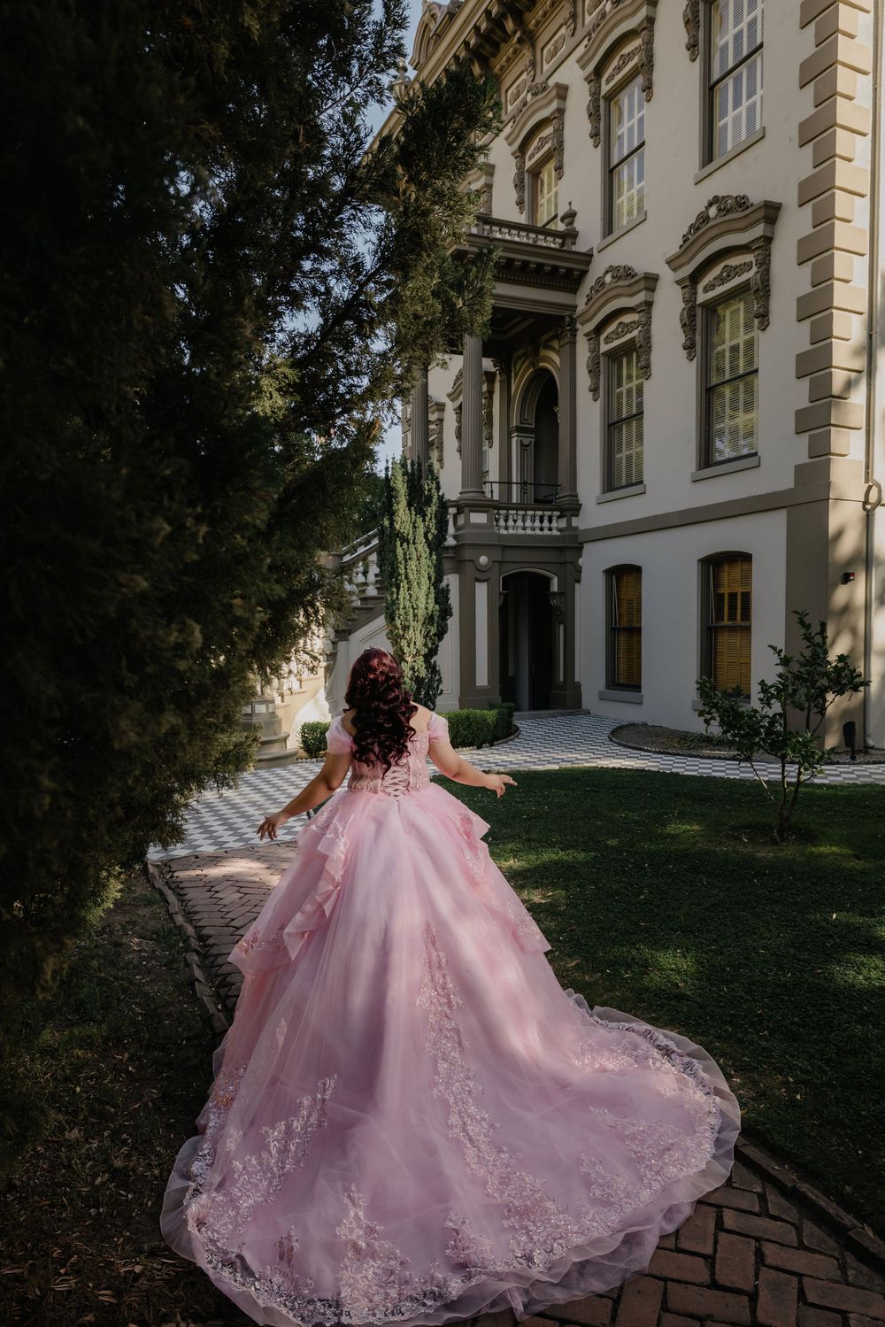 Young woman in a flowing pink ball gown with lace details walks toward an elegant mansion surrounded by lush greenery.