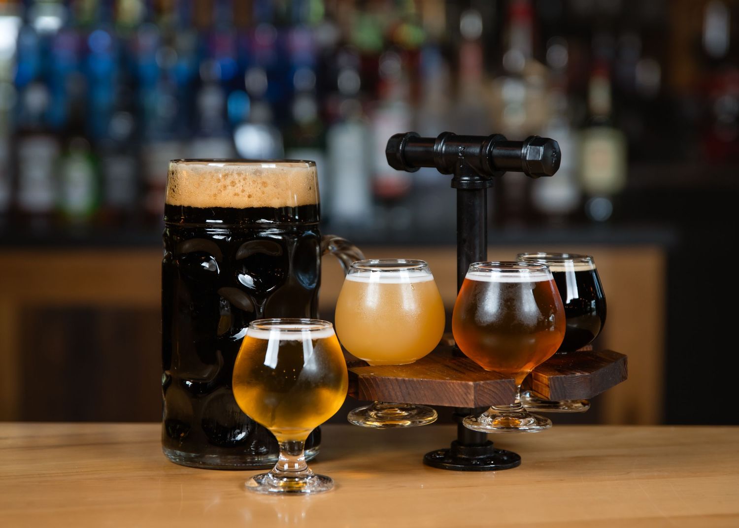 Assorted craft beers on a wooden bar, featuring a dark stout, golden ales, and a flight sampler with four varieties.
