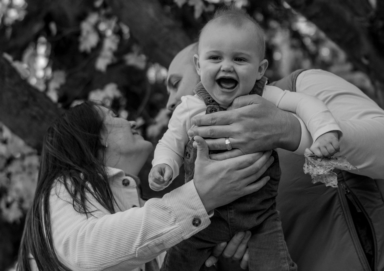 Joyful baby laughing while being lifted by parents during an outdoor black and white photo session.