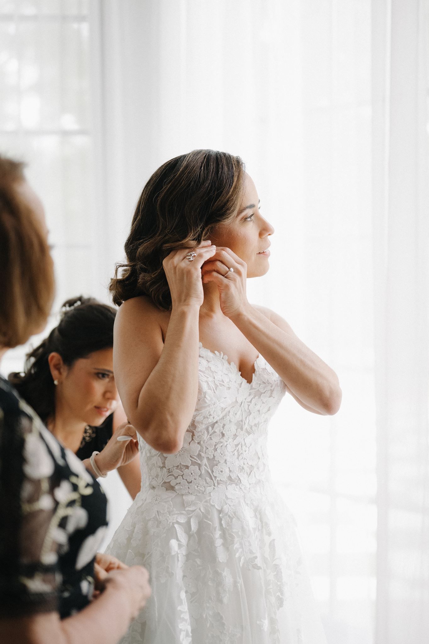 Bride in strapless lace wedding gown putting on earrings while bridesmaids help with dress in bright room.