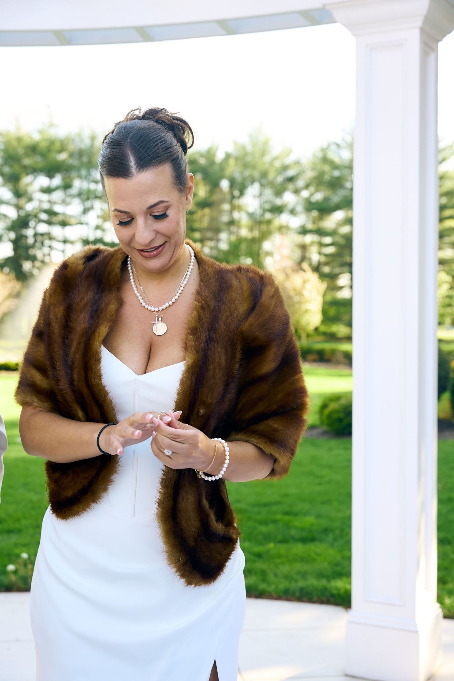 Bride in white dress, fur stole, and pearl jewelry adjusting bracelet outdoors near white columns.