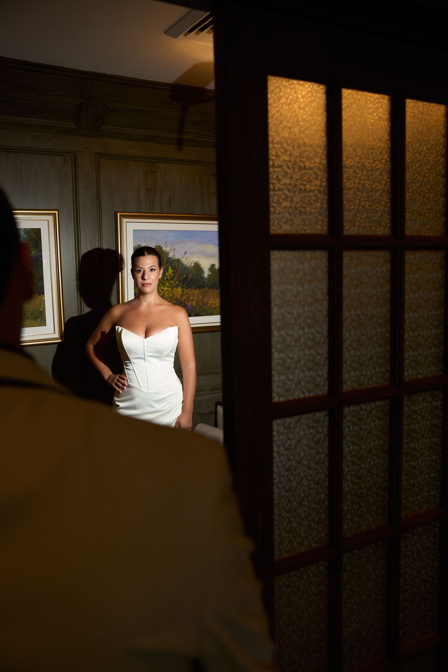 Bride in white strapless gown poses elegantly in a warmly lit room with framed artwork, viewed through a doorway.