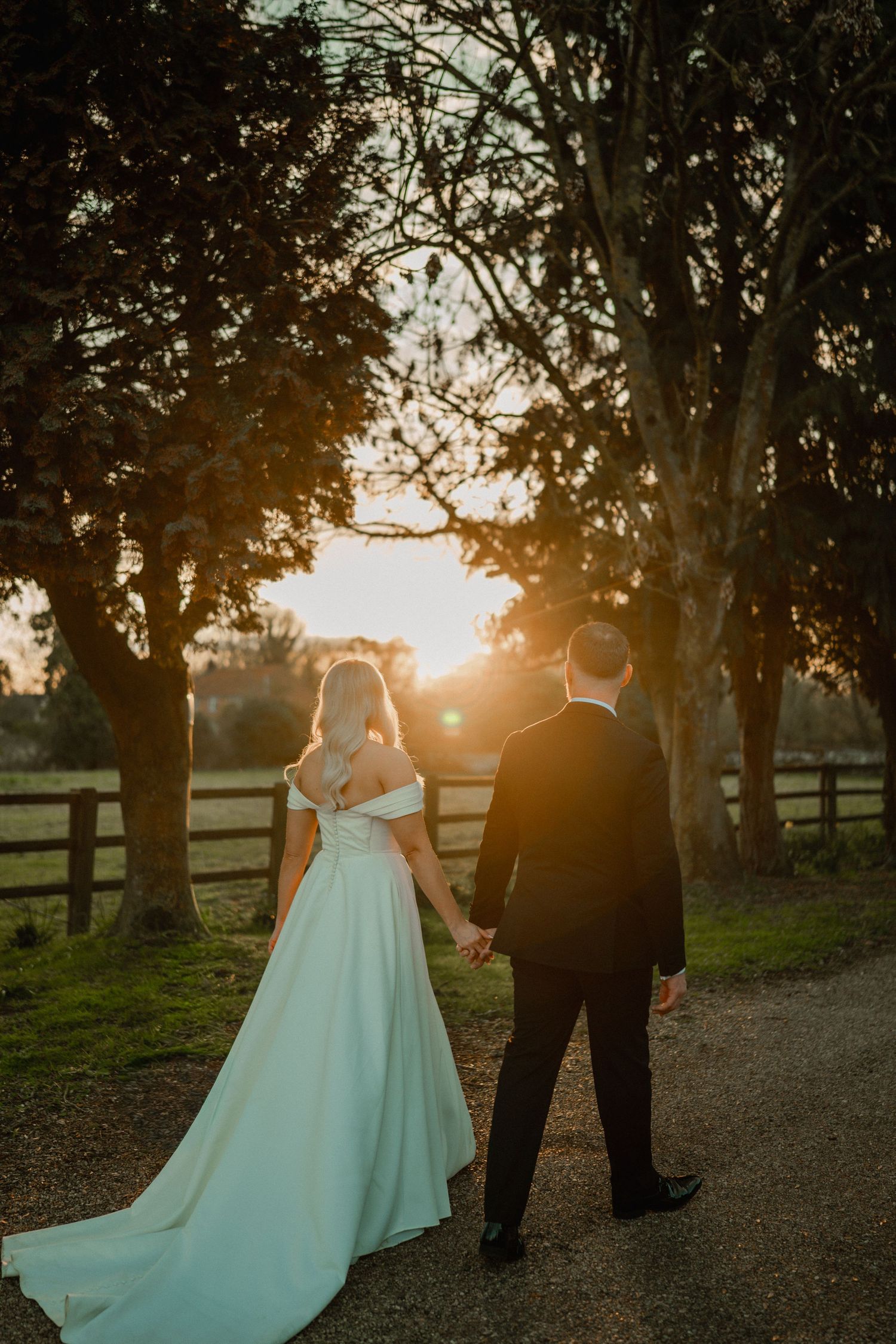Bride and groom holding hands walking toward golden sunset on a tree-lined path, bride in elegant white gown.